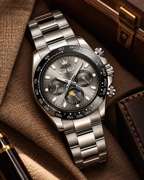 A stainless steel wristwatch with a silver-toned metal bracelet and a dark gray dial, styled on a brown textured fabric surface next to a leather pocket and wooden box.