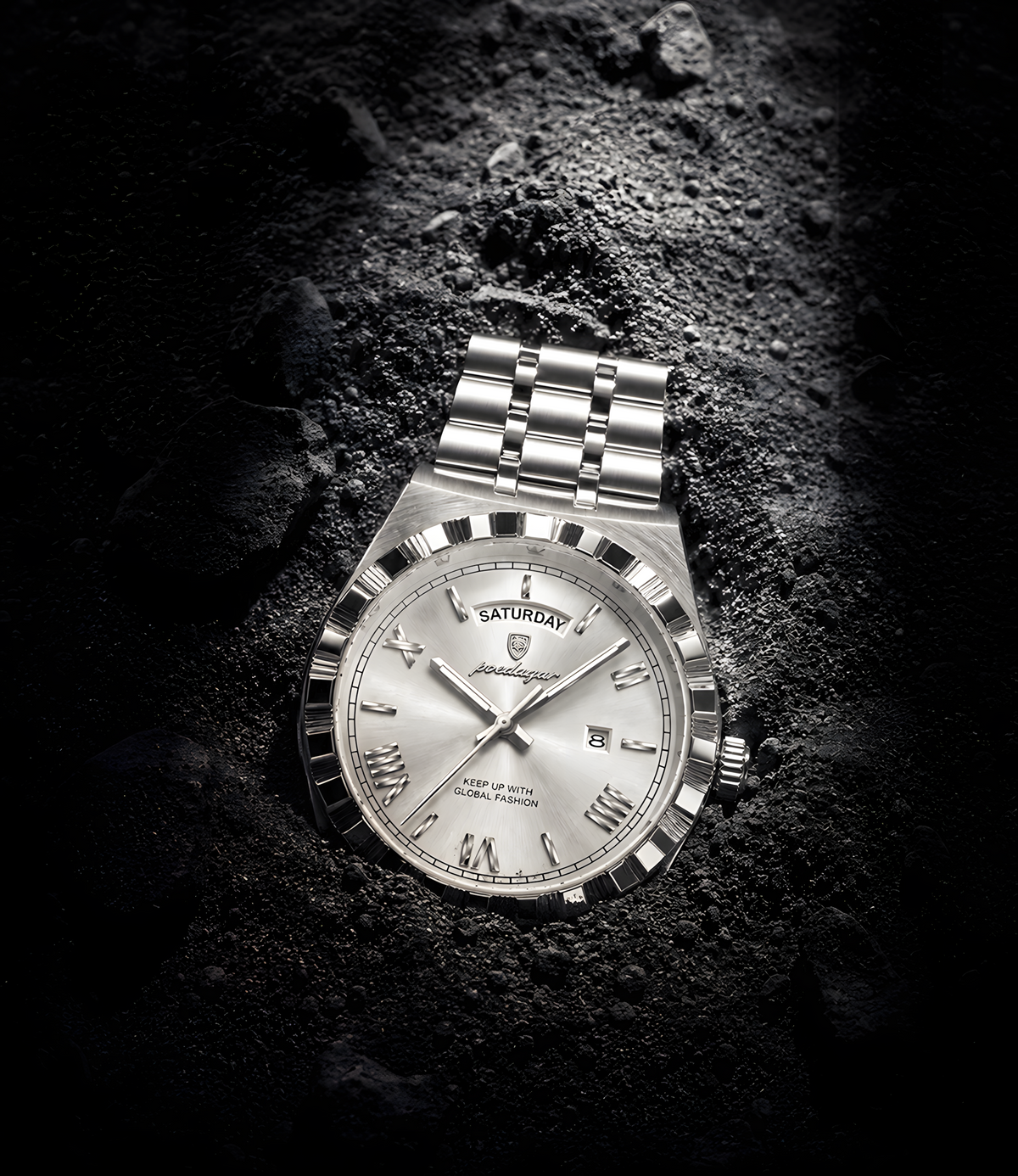 A silver-tone stainless steel wristwatch with a fluted bezel and matching link bracelet, resting on a dark textured rock surface. The watch features a silver dial with Roman numeral hour markers, a day/date window, and a shield logo on the dial.