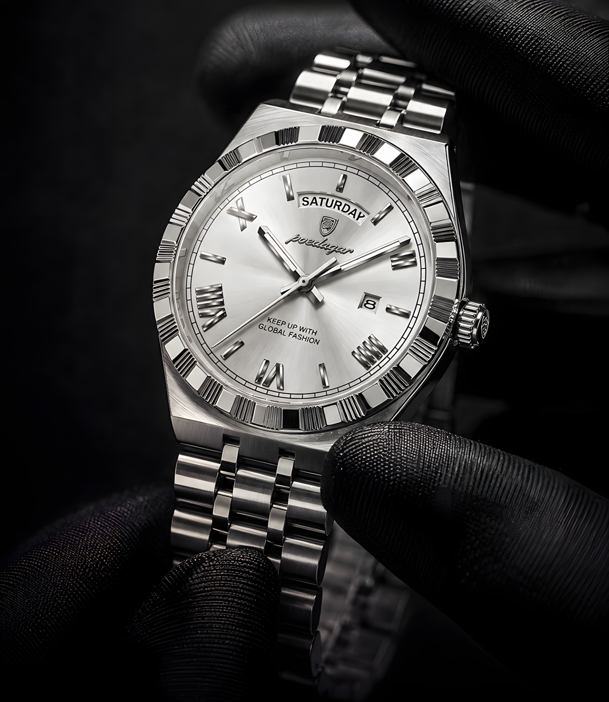 A silver stainless steel wristwatch with a white dial and Roman numeral hour markers, held in a gloved hand against a dark background. The watch features a date window showing Saturday and the brand name 'Judee' on the dial.